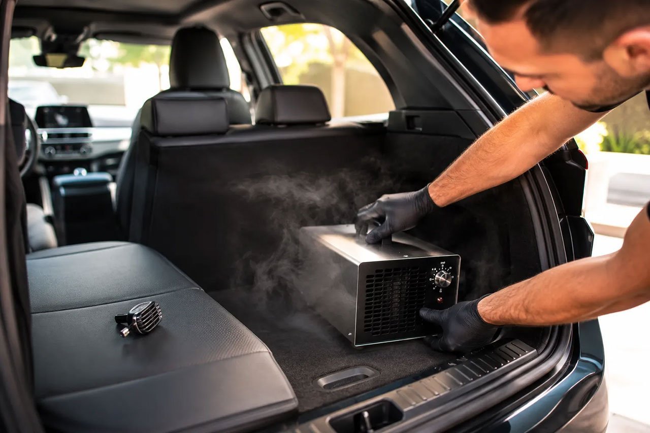 A professional detailer placing a compact ozone generator inside an open car trunk, warm interior lighting, steam-like ozone mist subtly visible, dashboard air freshener clip discarded on the seat beside it symbolizing comparison, bright daylight through windows, shallow depth of field, magazine automotive editorial style.